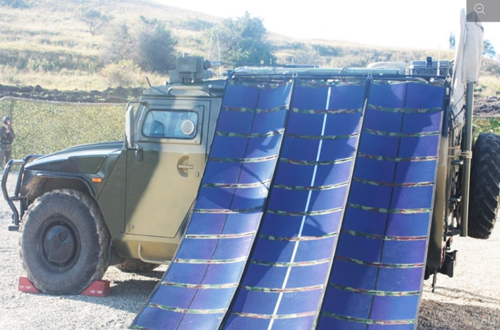 Желтый грейдер работает на подготовке участка под строительство солнечной электростанции — выравнивает красную глинистую почву на фоне зеленых холмов и леса.